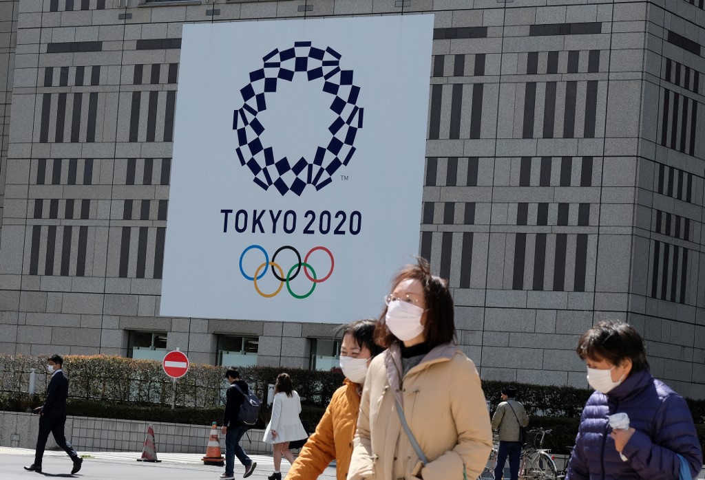 ON HOLD. Pedestrians wearing face mask walk before the logo of the Tokyo 2020 Olympic Games displayed on the Tokyo Metropolitan Government. Photo by Kazuhiro Nogi/AFP   