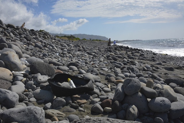 In this file photo, the remains of a car tire and a plastic bottle lay among the large stones as members of the media work in the background on the beach near Saint-Denis, where the officials recovered the metallic piece of debris, on the north coast of the Indian Ocean island of Reunion, August 2, 2015. Stringer/EPA  