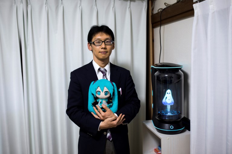 LOVE? In this photograph taken on November 10, 2018 Japanese Akihiko Kondo poses next to a hologram of Japanese virtual reality singer Hatsune Miku as he holds the doll version of her at his apartment in Tokyo, a week after marrying her. Photo by Behrouz Mehri/AFP 