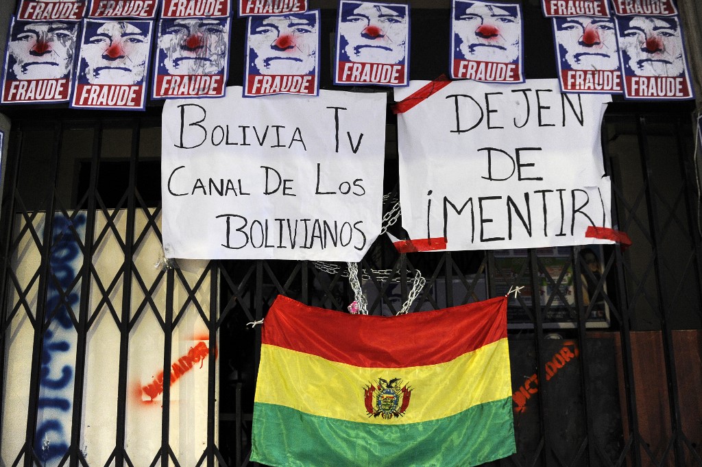 BOLIVIANS PROTEST. View of the closed entrance of the state TV channel BoliviaTV with pictures depicting Bolivian President Evo Morales as a clown reading "Fraud" and signs reading "Bolivia TV channel of Bolivians" and "Stop lying" next to a Bolivian national flag, after a protest on November 9, 2019, in La Paz. Photo by Jorge Bernal/AFP 
