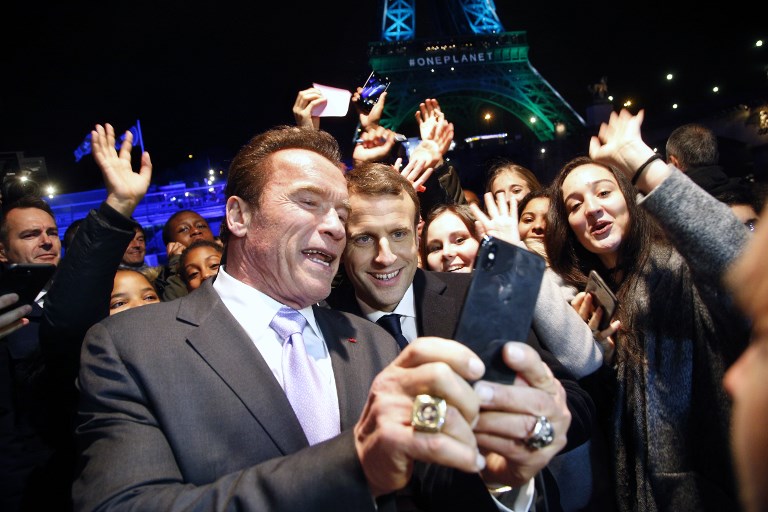 The TERMINATOR & THE PRESIDENT. Former Governor of California and US actor Arnold Schwarzenegger (L) and French President Emmanuel Macron (C) take a selfie with youths in front of the Eiffel Tower illuminated in green, aboard a boat cruising on the river Seine, after the One Planet Summit in Paris on December 12, 2017. Photo by Thibault Camus/AFP/POOL 